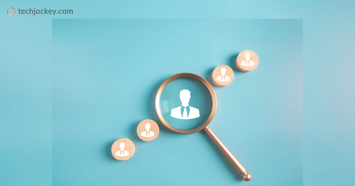 Magnifying glass highlighting a wooden icon of a person in a suit among several similar icons on a blue background, representing employee search or recruitment, with the techjockey.com logo in the top left corner.