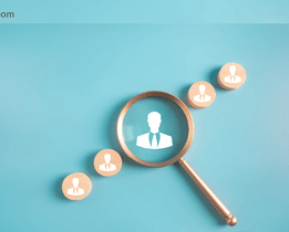 Magnifying glass highlighting a wooden icon of a person in a suit among several similar icons on a blue background, representing employee search or recruitment, with the techjockey.com logo in the top left corner.