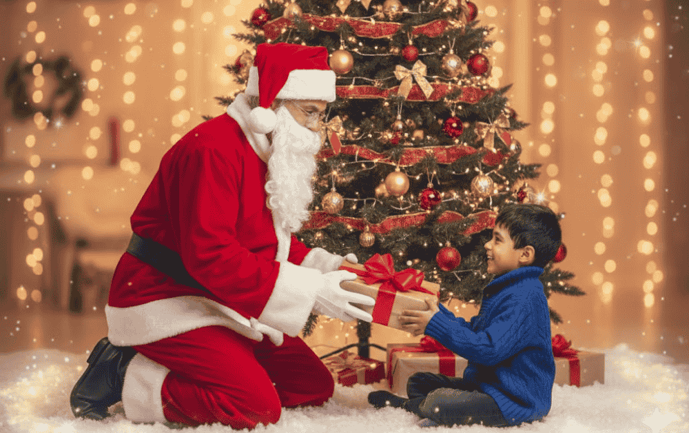 Santa Claus kneels by a decorated Christmas tree and hands a wrapped gift to a smiling young boy sitting in the snow.