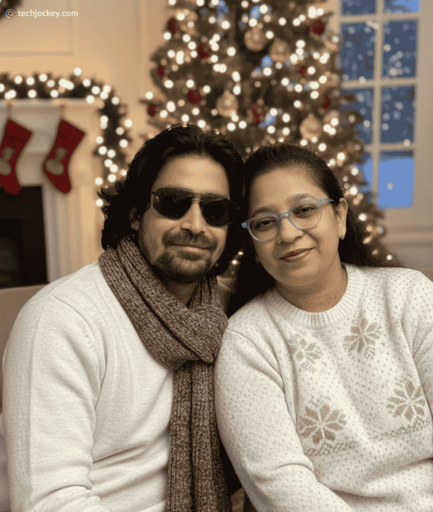 A couple dressed in cozy winter sweaters smiles while sitting in front of a decorated Christmas tree and festive lights.