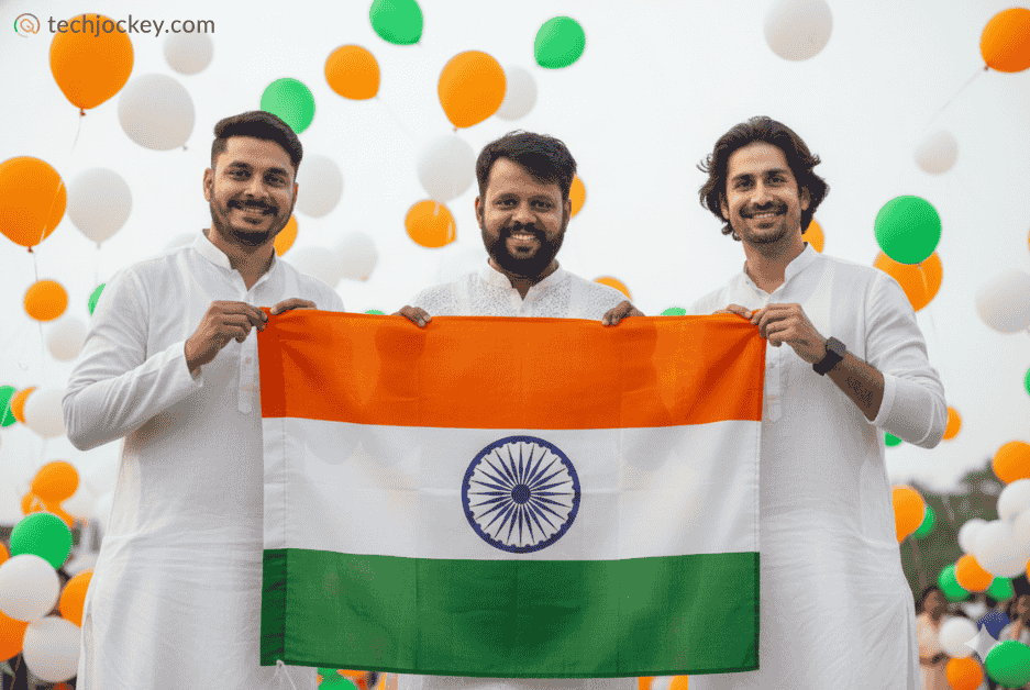Three men in traditional white attire holding the Indian national flag, celebrating Republic Day with tricolor balloons in the background.