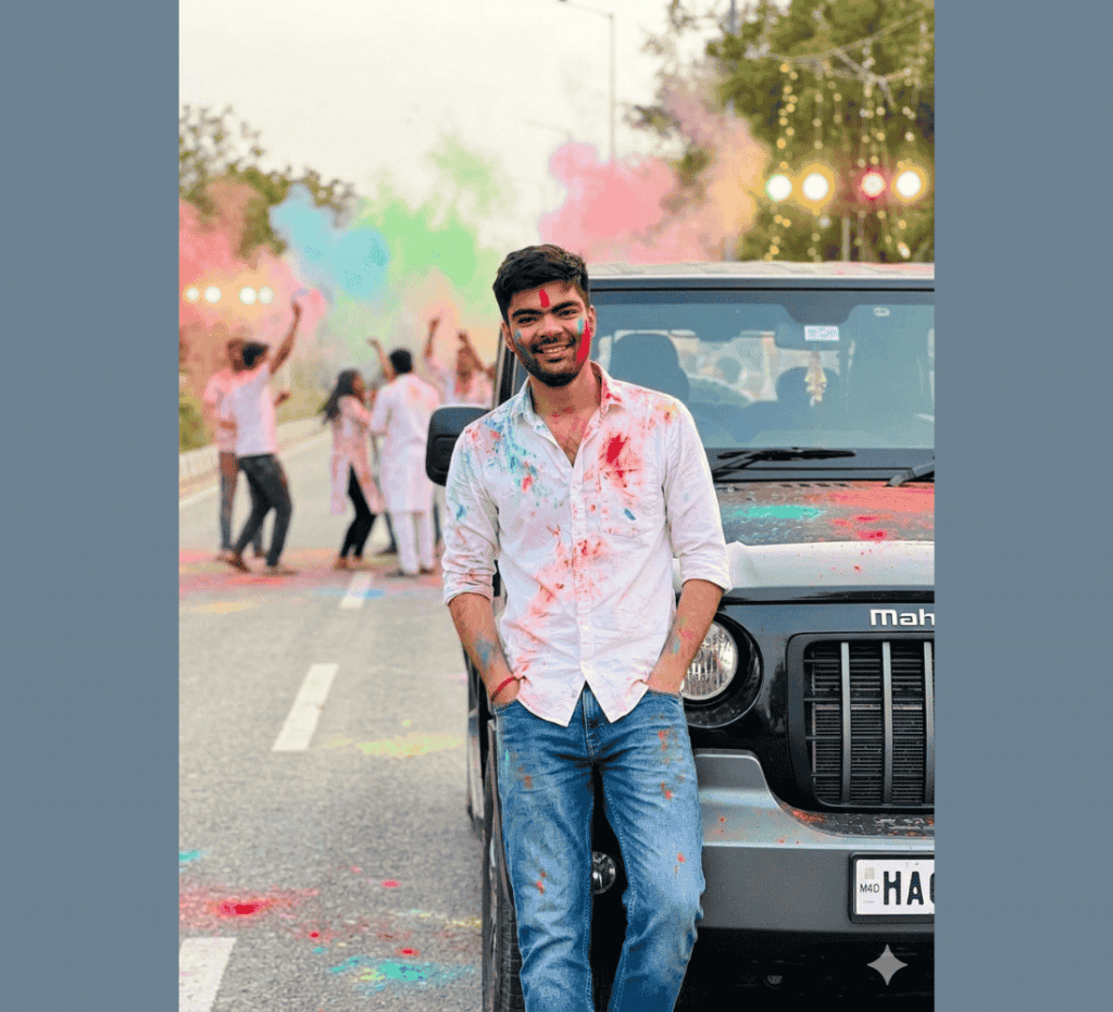 Young man leaning against SUV covered in Holi colors with friends celebrating in the background on street.
