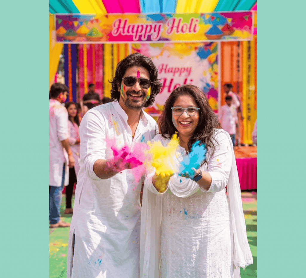 Man and woman holding colorful gulal under Happy Holi banner during festive celebration event.